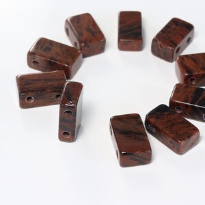 May include: Ten brown and black rectangular stone beads with two holes each. The beads are arranged in a circle on a white surface.