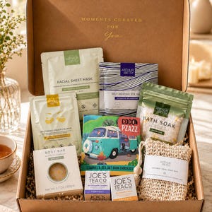 May include: A brown cardboard gift box filled with self-care products. The box contains facial sheet masks, eye masks, bath soak, a body bar, chocolate, tea, and a natural sisal soap bag. The box is labeled "Moments Curated For You."