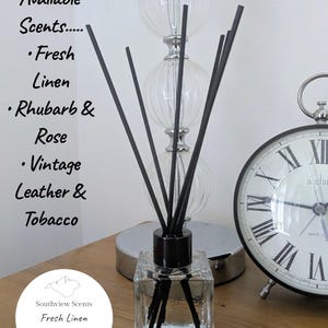May include: A clear glass reed diffuser with black reeds and a black cap. The available scents are Fresh Linen, Rhubarb & Rose, and Vintage Leather & Tobacco. The label reads "Fresh Linen" and "50ml." A decorative lamp and clock are in the background.