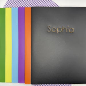 May include: A black notebook with the name "Sophia" in gold lettering. The notebook is next to a row of colorful paper sheets in green, yellow, blue, purple, and orange. The background is a purple and white checkered pattern.