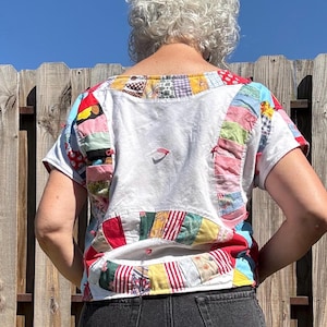 May include: A short-sleeved top constructed from a white base fabric featuring patchwork panels of various colorful patterned textiles. The garment includes a round neckline and a cropped silhouette, with visible stitching connecting the fabric pieces.