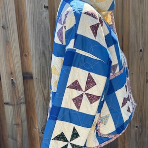 May include: A quilted jacket featuring a patchwork design of blue fabric borders and cream-colored squares. Each square contains a pinwheel pattern in various floral prints, including dark red, green, and yellow tones, with visible stitching.