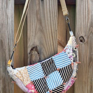 May include: A crescent-shaped crossbody bag made from a patchwork of various fabrics, including blue and white stripes, black and white stripes, and floral patterns. The bag features a beige canvas strap attached with metal swivel clasps.