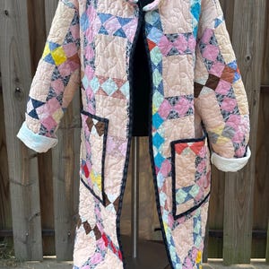 May include: A long-sleeved quilted coat featuring a patchwork design with various geometric patterns in shades of pink, blue, yellow, and brown. The coat has a stand-up collar, two large front patch pockets, and dark trim along the edges.
