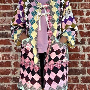 May include: A long-sleeved jacket constructed from a patchwork quilt featuring a diamond pattern in various colors including pink, yellow, purple, black, and white. The jacket has two front pockets and is secured at the chest with two white ties.