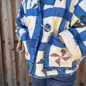May include: A quilted jacket featuring a patchwork design of blue, cream, and yellow fabric squares with pinwheel patterns. The jacket has a single large circular button closure, long sleeves with folded cuffs, and two front patch pockets.