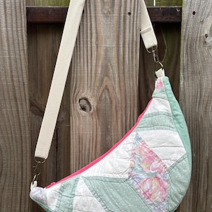 May include: A crescent-shaped crossbody bag constructed from quilted fabric featuring a patchwork design of sage green, white, and floral patterns in pink and blue. The bag includes a bright pink zipper closure and an adjustable beige canvas strap.