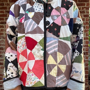 May include: A long-sleeved jacket constructed from a patchwork quilt featuring a variety of fabric patterns, including floral, plaid, and solid colors in shades of red, pink, black, white, and green. The jacket has a high collar and two black buttons.