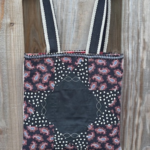 May include: A handmade tote bag with a black paisley pattern and a large black square in the center. The bag has black and white striped straps and is set against a wooden background. The bag's design includes red and blue accents.