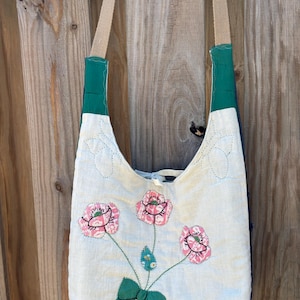 May include: Off-white shoulder bag with a floral design. The bag features three pink flowers with green stems and a green bow. The straps are tan and the top of the bag is trimmed with green fabric.