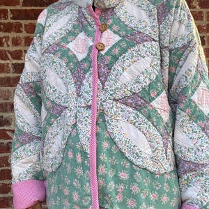 May include: A quilted jacket featuring a patchwork design of floral patterns in shades of green, white, and purple. The garment has a high collar, long sleeves with pink and white gingham cuffs, and a front closure with two wooden buttons and pink trim.
