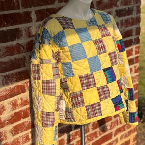 May include: A quilted jacket with a patchwork design. The jacket features a yellow base with squares of blue, red, and green plaid and solid fabrics. The jacket is displayed on a mannequin.