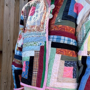 May include: A long-sleeve jacket constructed from a patchwork of various fabric scraps in diverse colors and patterns, including plaid, paisley, gingham, and solid tones. The jacket features a high collar, a front button closure, and pink trim.