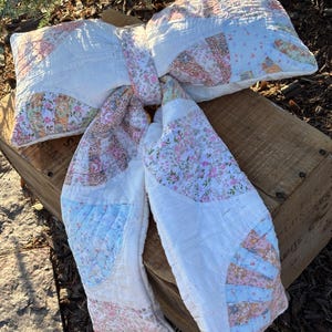 May include: A decorative bow-shaped pillow crafted from quilted fabric featuring a patchwork design. The textile displays a mix of white panels and sections with floral patterns in soft shades of pink, blue, and brown, finished with stitching.