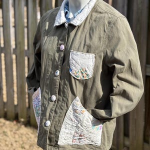 May include: Olive green jacket with white quilted accents on the pockets and cuffs. The jacket features a white collar and decorative buttons. The pockets and cuffs have colorful patchwork details, adding a unique, handcrafted touch to the garment.
