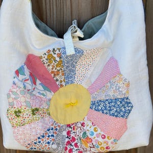May include: White fabric shoulder bag with a large, colorful, patchwork flower design. The flower features various patterned fabric petals in pink, blue, and floral prints, with a yellow center. The bag has a drawstring closure and yellow embroidered details.