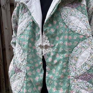 May include: A quilted jacket featuring a patchwork design of green, white, and pink floral fabric patterns. The garment has a wide collar, a white interior lining, and is fastened at the center with a decorative silver metal clasp over white lace.