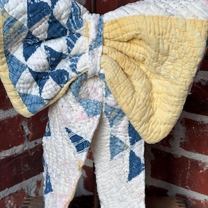 May include: A decorative bow crafted from quilted fabric featuring a patchwork pattern of blue and white triangles on one side and solid pale yellow on the other. The fabric has a visible stitched texture and hangs from a central metal stand.