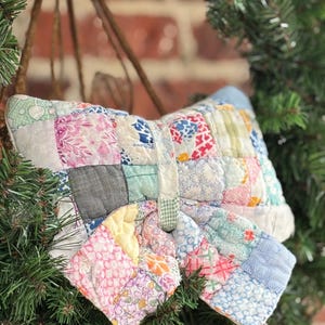 May include: A handmade, vintage-style quilted pillow with a patchwork design. The pillow features a variety of fabric patterns in pastel colors, including pink, blue, and yellow. The pillow is displayed on a Christmas wreath.