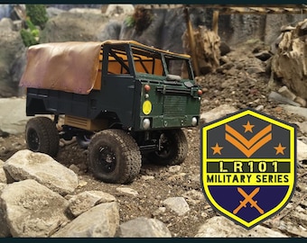 Op de afbeelding: Een donkergroen militair vrachtwagenmodel met een bruin canvas zeil, geplaatst op een rotsachtig terrein. De vrachtwagen heeft grote, zwarte banden en een gele koplamp. Een badge met de tekst "LR101 MILITARY SERIES" is zichtbaar.