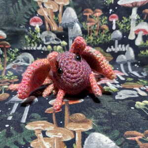 May include: A small, handmade crochet creature featuring a round, dark purple head with two black plastic eyes. The body is crafted from variegated yarn in shades of pink, orange, and purple, with large, wide-set ears and several small, dangling legs.