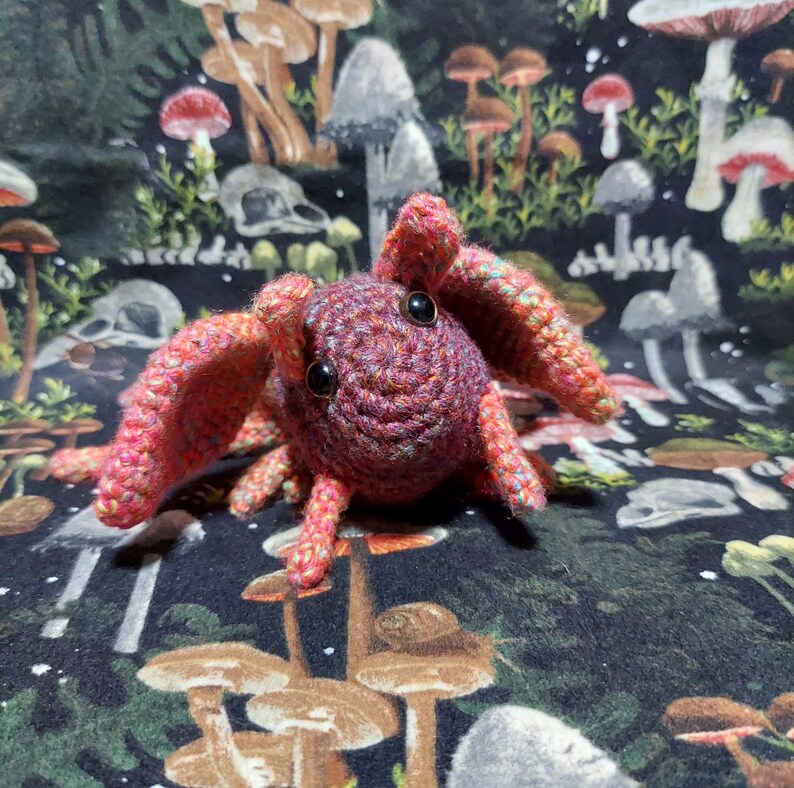 May include: A small, handmade crochet creature featuring a round, dark purple head with two black plastic eyes. The body is crafted from variegated yarn in shades of pink, orange, and purple, with large, wide-set ears and several small, dangling legs.