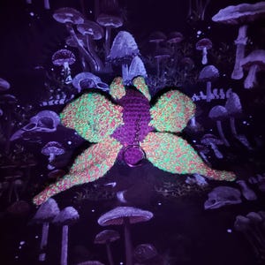 May include: A crochet moth keychain featuring a dark purple central body and four large, textured wings in a vibrant neon green and pink variegated yarn. A silver-toned metal split ring is attached to the center of the moth's back for hanging.