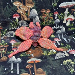 May include: A crocheted butterfly keychain featuring a purple central body and four large, textured wings in a variegated blend of orange, pink, and yellow yarn. A silver-toned metal split ring is attached to the center of the butterfly body.