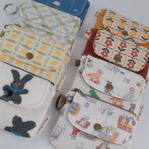 May include: A collection of small, patterned fabric pouches with key rings. Designs include blue denim with bows, floral patterns, and animal illustrations. Each pouch has a snap closure and a small pocket.