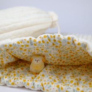 Puede incluir: Una pequeña figurita de animal de cerámica amarilla con forma de oveja asoma de una bolsa de cordón blanco con estampado floral amarillo.