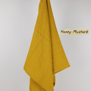 Puede incluir: Un pa&ntilde;o de cocina de color amarillo mostaza miel colgado de una rama de madera. El pa&ntilde;o tiene una peque&ntilde;a etiqueta blanca en la esquina inferior con un estampado gr&aacute;fico negro. El texto 'Honey Mustard' est&aacute; escrito en negro a la derecha.