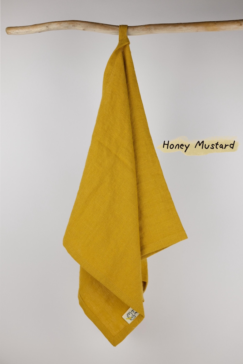 Puede incluir: Un pa&ntilde;o de cocina de color amarillo mostaza miel colgado de una rama de madera. El pa&ntilde;o tiene una peque&ntilde;a etiqueta blanca en la esquina inferior con un estampado gr&aacute;fico negro. El texto 'Honey Mustard' est&aacute; escrito en negro a la derecha.