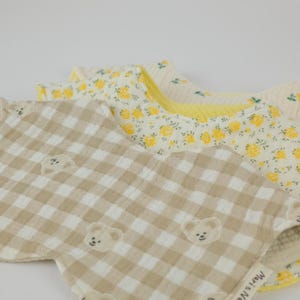 Pu&ograve; includere: Due bavaglini in tessuto arrotondati sovrapposti. Il bavaglino superiore presenta un motivo a quadretti vichy beige e bianco con piccoli volti di orso e un'etichetta con la scritta Mari & Nari. Quello inferiore ha uno sfondo bianco con fiori gialli.