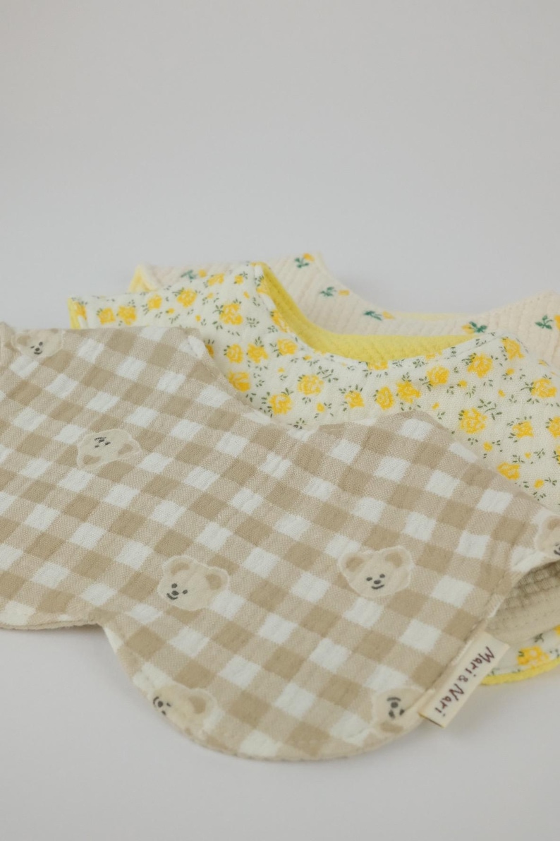 Pu&ograve; includere: Due bavaglini in tessuto arrotondati sovrapposti. Il bavaglino superiore presenta un motivo a quadretti vichy beige e bianco con piccoli volti di orso e un'etichetta con la scritta Mari & Nari. Quello inferiore ha uno sfondo bianco con fiori gialli.