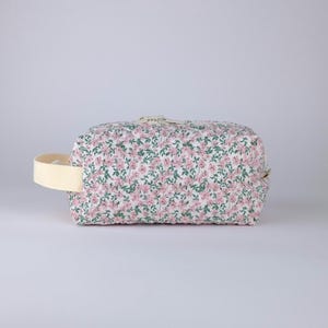 May include: A rectangular cosmetic bag with a floral print. The bag has a white background with a pattern of pink flowers and green leaves. It has a cream-coloured handle and a zip closure.