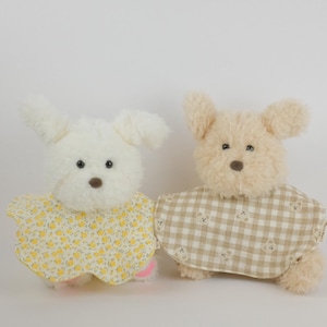 Pu&ograve; includere: Due peluche a forma di cane con pelliccia soffice. Il cane a sinistra &egrave; bianco e indossa una mantellina gialla con un piccolo motivo floreale. Il cane a destra &egrave; beige e indossa una mantellina a quadri beige e bianchi con piccoli orsi.
