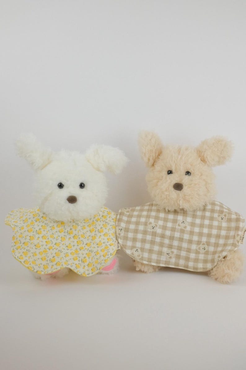 Pu&ograve; includere: Due peluche a forma di cane con pelliccia soffice. Il cane a sinistra &egrave; bianco e indossa una mantellina gialla con un piccolo motivo floreale. Il cane a destra &egrave; beige e indossa una mantellina a quadri beige e bianchi con piccoli orsi.
