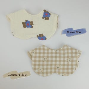 Pu&ograve; includere: Due bavaglini per neonati con bordi smerlati. Il bavaglino superiore &egrave; color crema con una stampa di orsi marroni che indossano magliette blu. Il bavaglino inferiore presenta un motivo a quadri beige e bianco con piccoli musi d'orso. Etichetta Meri&Nuri.