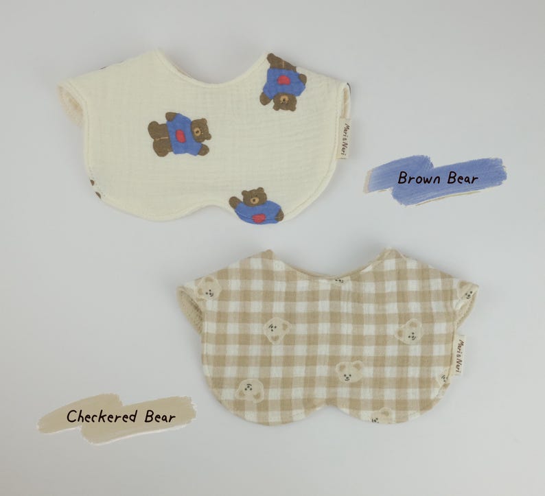 Pu&ograve; includere: Due bavaglini per neonati con bordi smerlati. Il bavaglino superiore &egrave; color crema con una stampa di orsi marroni che indossano magliette blu. Il bavaglino inferiore presenta un motivo a quadri beige e bianco con piccoli musi d'orso. Etichetta Meri&Nuri.
