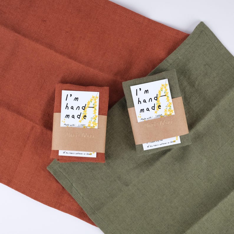 May include: Two linen tea towels, one rust orange and one olive green, with a small tag attached to each. The tag is brown with white text that reads "I'm hand-made with linen" and "Marte Neri".