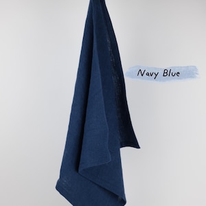 Puede incluir: Un pa&ntilde;o de cocina azul marino con una textura tejida visible, colgado de una rama de madera clara. La tela est&aacute; doblada, mostrando un borde con dobladillo y un peque&ntilde;o lazo en la parte superior para colgar. El texto Navy Blue est&aacute; a la derecha.