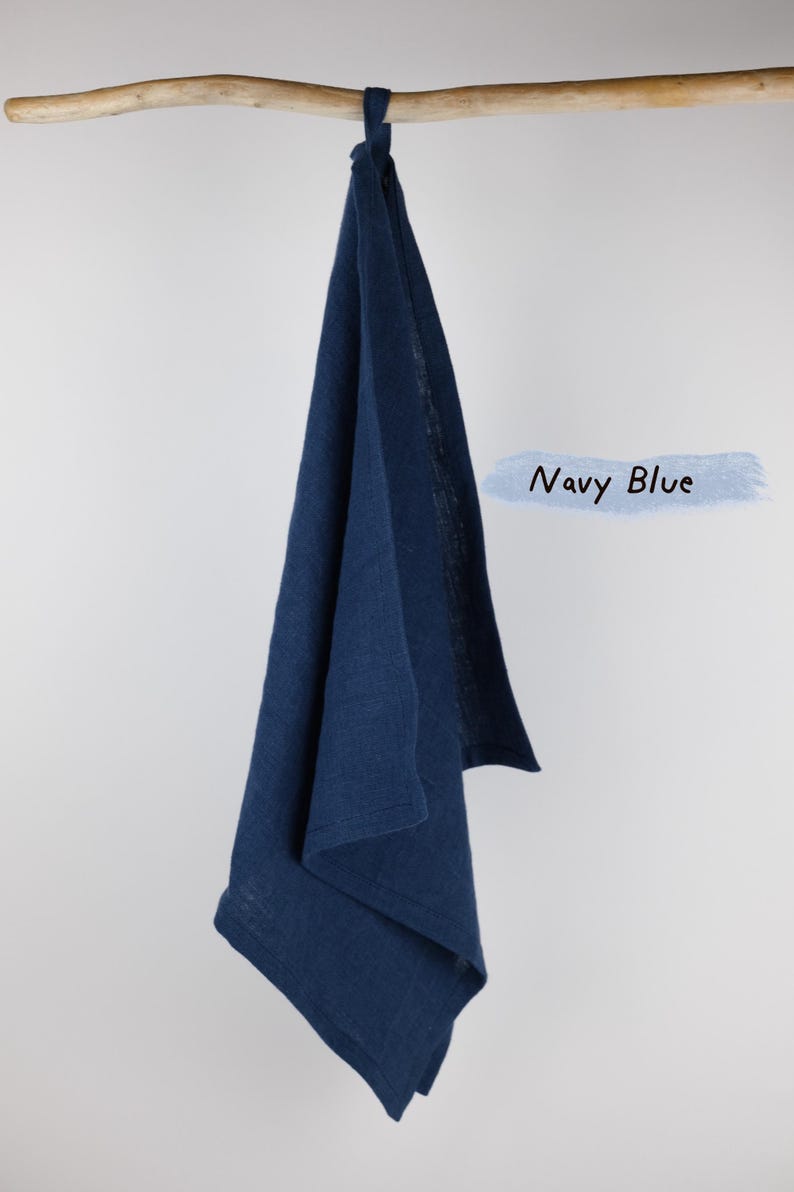 Puede incluir: Un pa&ntilde;o de cocina azul marino con una textura tejida visible, colgado de una rama de madera clara. La tela est&aacute; doblada, mostrando un borde con dobladillo y un peque&ntilde;o lazo en la parte superior para colgar. El texto Navy Blue est&aacute; a la derecha.