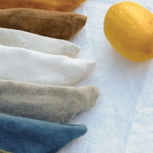 Puede incluir: Una colecci&oacute;n de servilletas de tela dobladas en varios colores, incluyendo &oacute;xido, amarillo mostaza, marr&oacute;n, beige, blanco, gris, azul y verde oliva, apiladas junto a un lim&oacute;n amarillo brillante sobre una superficie de tela blanca texturizada.
