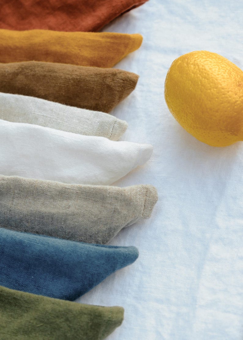 Puede incluir: Una colecci&oacute;n de servilletas de tela dobladas en varios colores, incluyendo &oacute;xido, amarillo mostaza, marr&oacute;n, beige, blanco, gris, azul y verde oliva, apiladas junto a un lim&oacute;n amarillo brillante sobre una superficie de tela blanca texturizada.