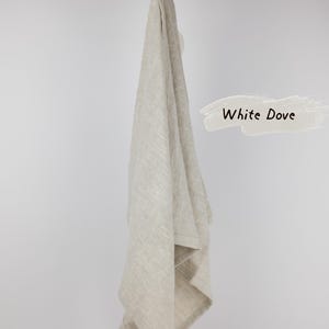 Puede incluir: Un pa&ntilde;o de cocina de lino beige con un lazo para colgar, drapeado sobre una rama de madera. La tela tiene una textura tejida visible. A la derecha, un gr&aacute;fico de pincelada clara contiene el texto negro White Dove, que identifica el color.