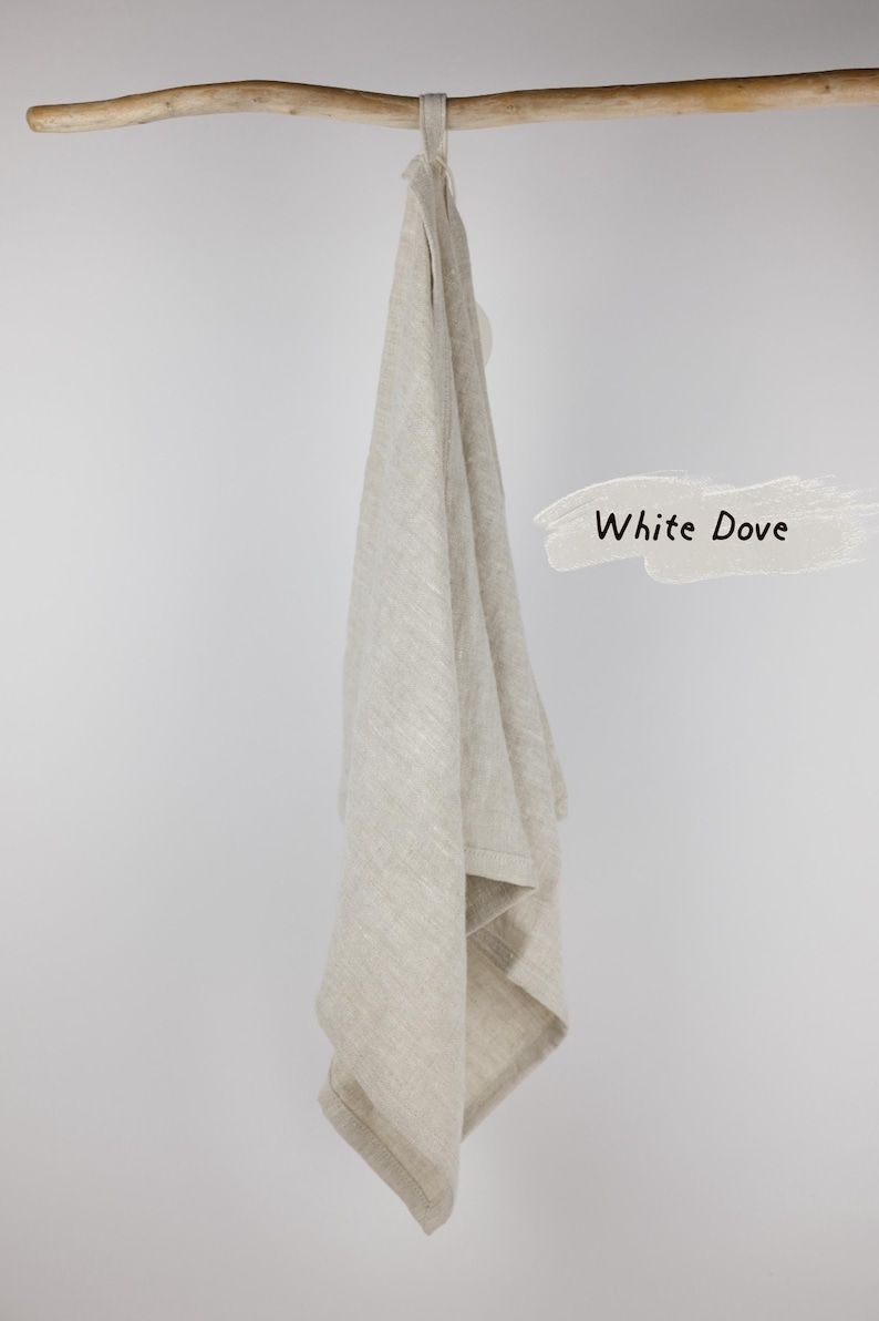 Puede incluir: Un pa&ntilde;o de cocina de lino beige con un lazo para colgar, drapeado sobre una rama de madera. La tela tiene una textura tejida visible. A la derecha, un gr&aacute;fico de pincelada clara contiene el texto negro White Dove, que identifica el color.