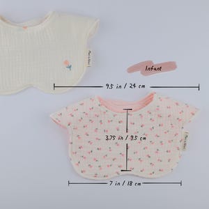 Pu&ograve; includere: Due bavaglini per neonati in tessuto testurizzato. Uno &egrave; color crema con un piccolo fiore ricamato e una larghezza di 24 cm. L'altro presenta un motivo floreale rosa, misura 18 cm di larghezza e 9,5 cm dal collo al bordo inferiore.