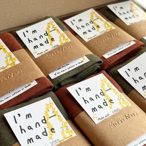 May include: A stack of folded linen tea towels with brown paper packaging. Each towel has a label that reads "I'm hand made" and "Made with: linen". The label also includes the name "Maria Nari".