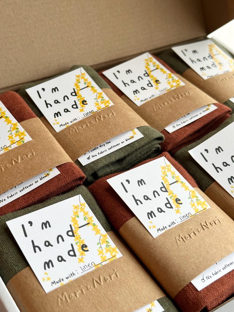 May include: A stack of folded linen tea towels with brown paper packaging. Each towel has a label that reads "I'm hand made" and "Made with: linen". The label also includes the name "Maria Nari".