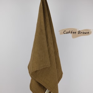 Puede incluir: Un pa&ntilde;o de cocina color marr&oacute;n caf&eacute; con una textura tejida visible que cuelga de una vara de madera. La tela tiene una peque&ntilde;a etiqueta cosida en la esquina inferior. El texto Coffee Brown est&aacute; impreso en una fuente cursiva oscura a la derecha.